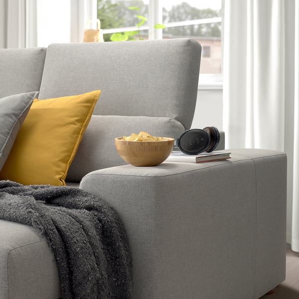 Comfortable grey ISTABY sofa, armrest with snacks, headphones, book, yellow pillow, cozy blanket, large window.