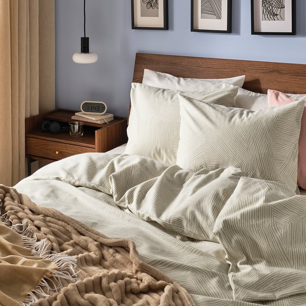 Cosy bedroom with beige JÄRNTRÄD duvet, wooden headboard, pink pillow, and bedside table with lamp, clock, and books.