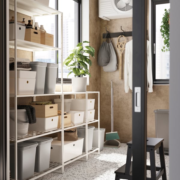 Organized storage room with white shelves holding containers, boxes, and plants on a tiled floor, with wall hangings. Length: 149 characters