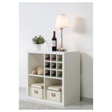 A white KALLAX shelving unit holding wine bottles, storage boxes, and a lamp.