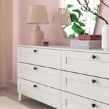 White dresser with six drawers, black knobs, lamp, and books.