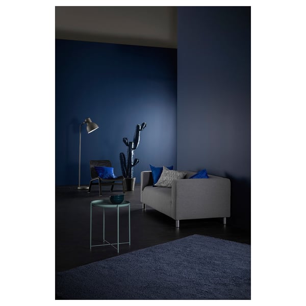 Modern living room with grey KLIPPAN sofa, blue cushions, side table, lamp, and cactus. Dark blue walls, carpet, and dramatic lighting.