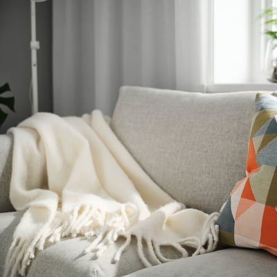 Off-white cozy throw blanket with fringe, larger than cushion, on cream sofa.