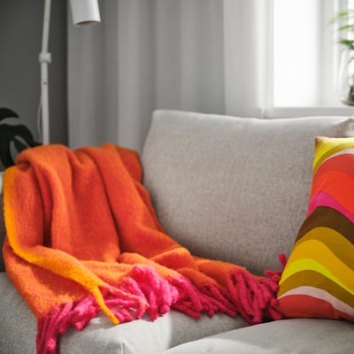Bright pink/orange KLIPPOXEL throw blanket on sofa, soft and fluffy, with fringe ends, near large window, covers part of grey cushion.