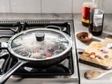 A covered frying pan on a stove. Nearby, two bottles and chopped vegetables on a wooden board.