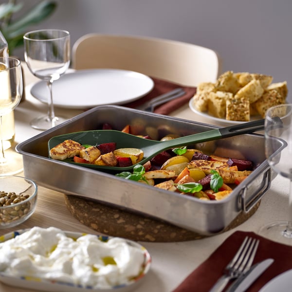 A KNORRHANE green kitchen utensil in a pan with roasted vegetables, beside white cream, bread, plates, glasses, and cutlery on a light-colored table.