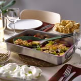 A KNORRHANE green kitchen utensil in a pan with roasted vegetables, beside white cream, bread, plates, glasses, and cutlery on a light-colored table.