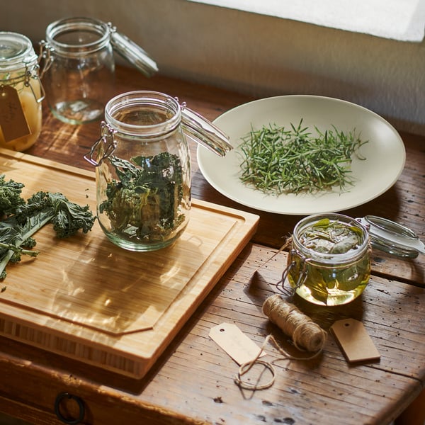 Wooden cutting board with jars filled with herbs, one with metal handle, beside a plate of rosemary, surrounded by jars and twine.