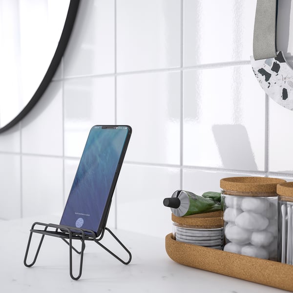 On a marble surface, a black metal phone stand holds a smartphone at an angle next to cork jars with office supplies.