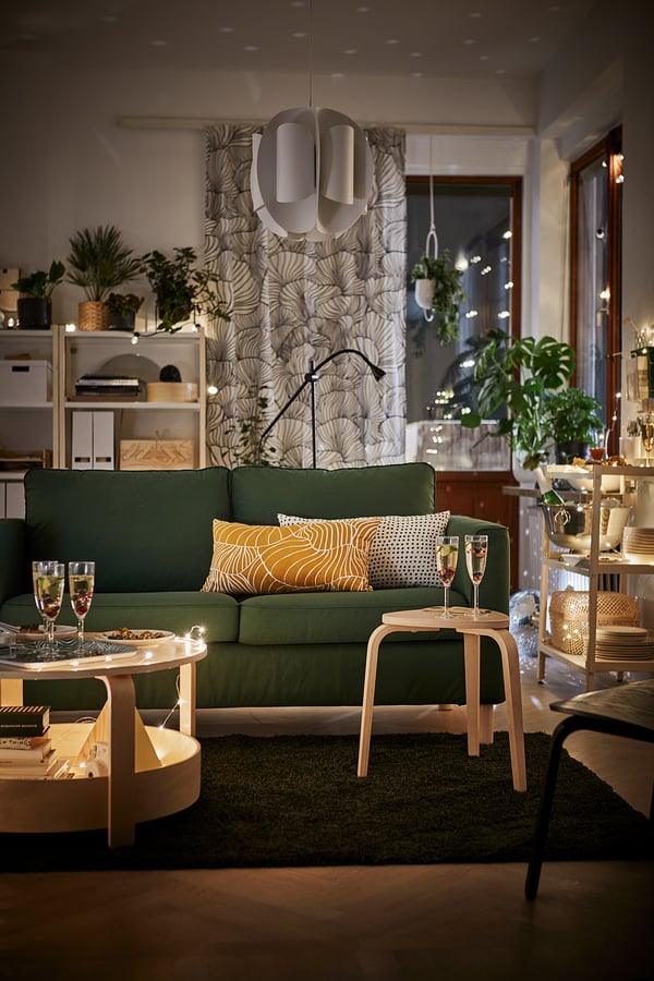 Modern living room with KYRRE birch wood stools, green sofa, yellow pillows, white pendant lamp, and festive decorations including champagne glasses.