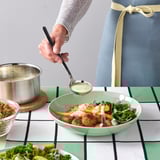 A person pours creamy sauce onto a green bowl filled with fish, grains, and vegetables.