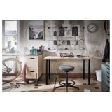 Industrial-style workspace with LAGKAPTEN desk, chair, tools, and decorative items. Concrete wall, tiled floor, and large window.