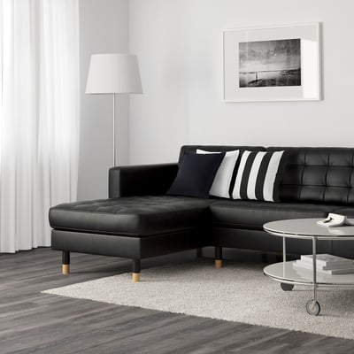Modern living room with black l-couch, striped/solid pillows, white lamp, book table, large rug on dark wood floor.