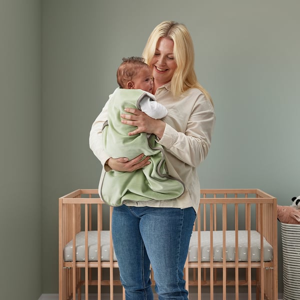 Woman smiles holding baby in green sleeping bag between cribs.