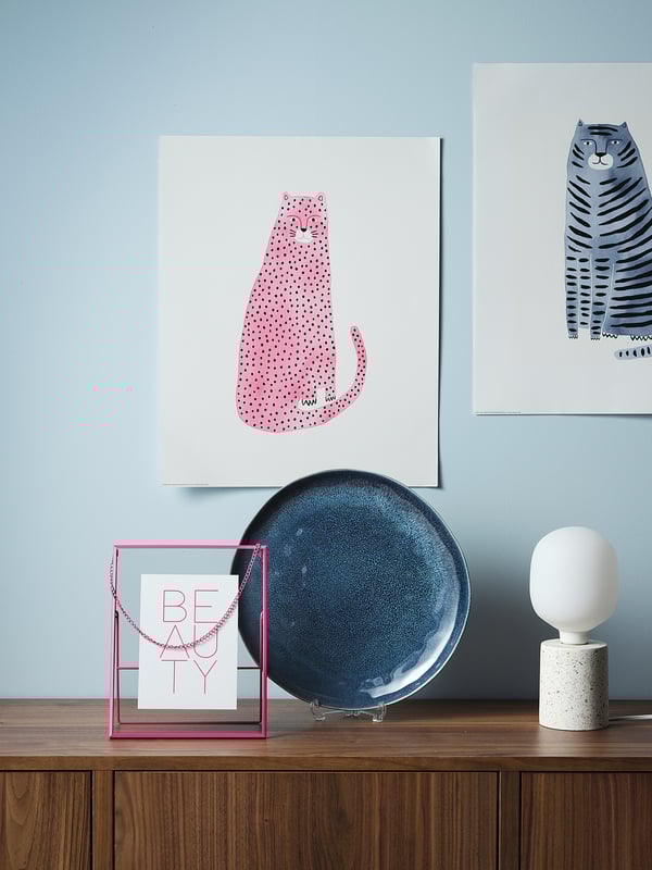 A stylish display on a dark wood shelf: colorful cat prints, blue plate, and a white terrazzo-table lamp.