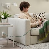 Person knitting on beige couch with a green LILLHULT usb cable. Couch has a patterned pillow and green blanket.