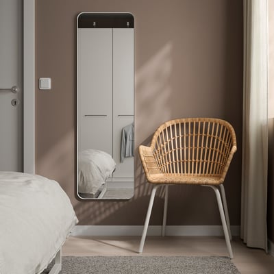 A LINDBYN mirror with white frame, rounded corners, reflecting a bed and wardrobe. Beside it, a white woven chair.