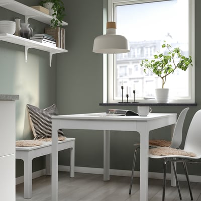 Cozy workspace with white table, two cushioned chairs, green walls, lamp & plant.