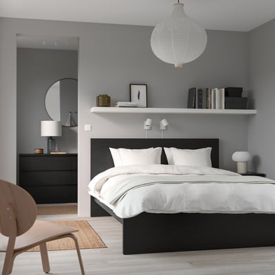 Modern minimalist bedroom with large bed, grey walls, white bedding, floating shelves with books, mirror, light wood floors, and pendant light.