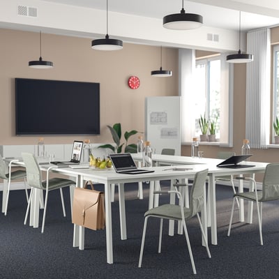 Modern office with white tables, laptops, grey chairs, lights, plants, and large screen.