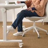 Person sitting at adjustable table with footrest, taking notes. White sneakers, jeans, and sweater visible.