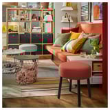 Bright living room with orange sofa, colorful pillows, striped rug, wire furniture, and plants. Centered orange stool.
