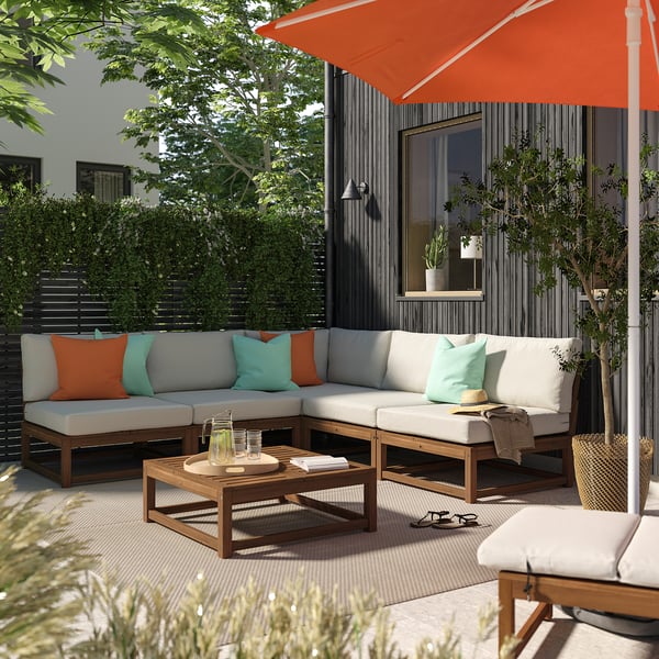 Modern patio with sectional sofa, orange accents, coffee table, umbrella, lush greenery.