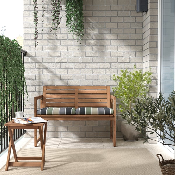 Modern outdoor patio with light brown acacia wood bench, striped cushions, side table, and potted plants against gray brick wall.