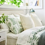 Cosy bed with green-flowered white NÅLBJÖRNBÄR duvet, cream pillows, plants, and green lamp.