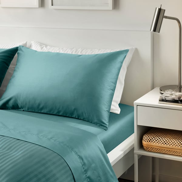 Green-blue nattajjasmin bed set; smooth, sateen-woven cotton-lyocell, rectangular pillows on a white bed, beside small, modern bedside table.