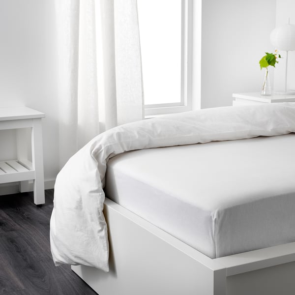 Neatly made bed with white sheets, lamp, and greenery beside it, large windows with sheer curtains.