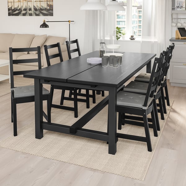 Black NORDVIKEN dining set with extendable table and 6 chairs. Modern-rustic, seats 8. Cozy, light-filled room with couch and lamp.