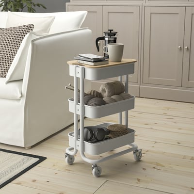 Small white rolling cart, perfect for storage, fits near sofa. Top has wooden lid, bottom has bins. Holds mugs, books, baskets.