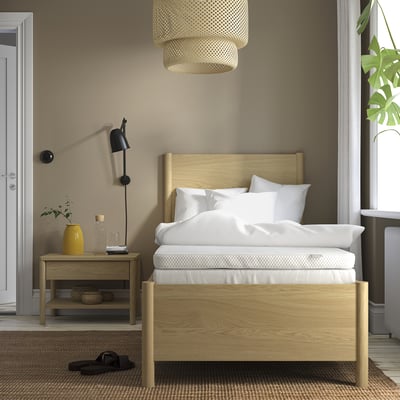 Minimalist bedroom: light wood bed, white linens, small bedside table. Bright light fixture, plant by window, cosy rug.
