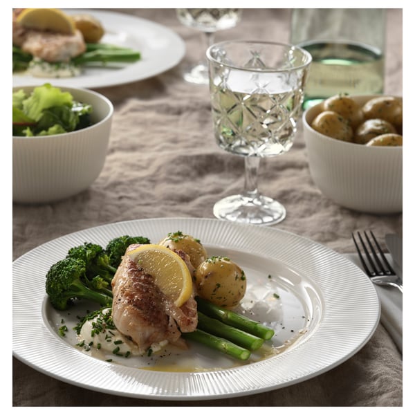 White dinner plate with lemon chicken, broccoli, and asparagus. Surrounded by bowls, a wine glass, and cutlery.