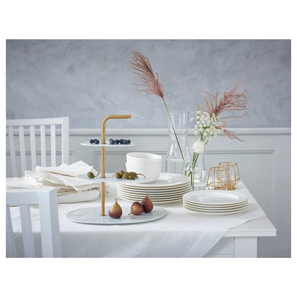Elegant white dinner setup on table. Includes stacked plates, tiered stand with fruits, and gold candle holder with dried flowers in vase.
