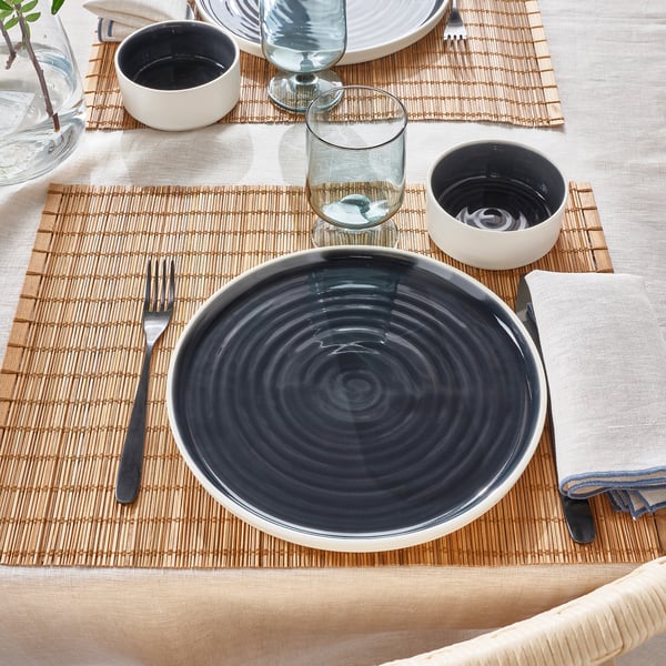 Table setting featuring minimalist OMBONAD dishes. Includes a ceramic plate, small bowls, glass, fork, and napkin on a woven mat.