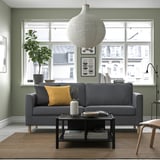 Modern living room with grey sofa, yellow pillow, large window, sage walls, plant, coffee table books.