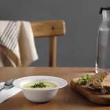 Bowl of soup and bread on wooden table. Tempered glass bowl with decorative rim and green soup garnished with herbs.