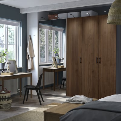 Modern bedroom with large PAX wardrobe in wooden colour, beside a desk and black chair, featuring a mirror and woven textiles.