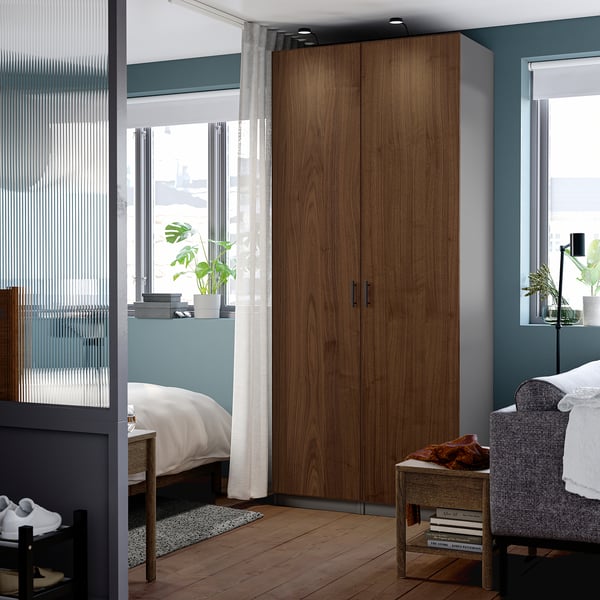 Modern bedroom with tall wooden wardrobe. Soft curtains by windows, white bed, gray chair. Wardrobe is large, fits room well.