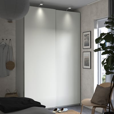 Modern bedroom with white PAX wardrobe, black bed, wooden chair, and green plant beside a large window.