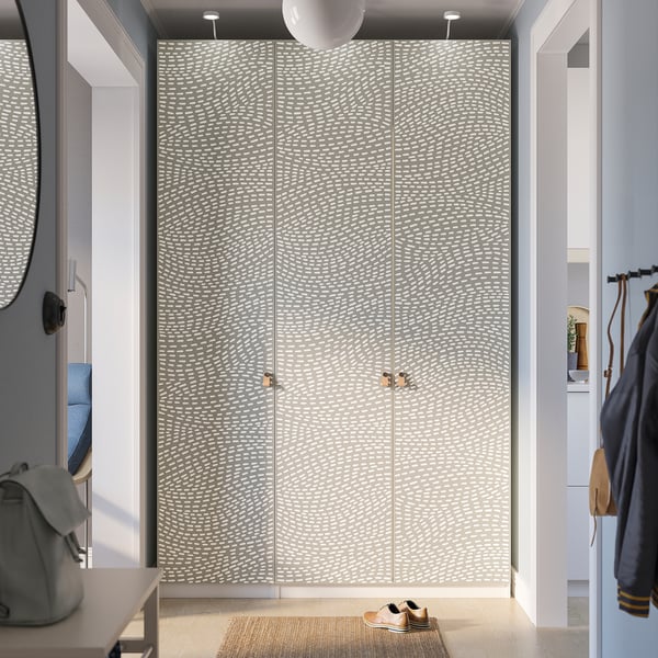 A modern wardrobe named PAX in a hallway. It white and gray wavy-patterned doors with gold handles. It stands tall next to a coat hanger and a mirror.