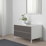 Modern PLATSA storage with 2 grey drawers, white top, plant, books, against white wall.