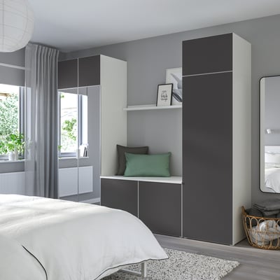Modern bedroom with gray PLATSA cabinets, mirrored doors, and bench. Green & black cushions, windows with gray curtains, white bed, & woven basket.