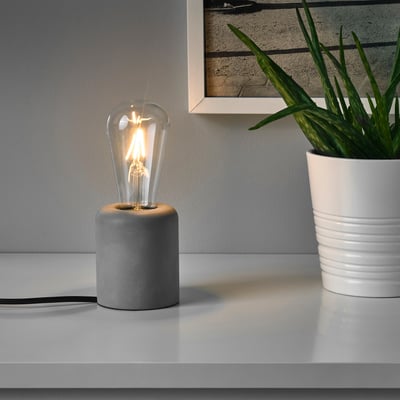 A small table lamp with a concrete base sits on a white surface, its glowing bulb providing warm, soft light. Beside it, a potted plant adds greenery.