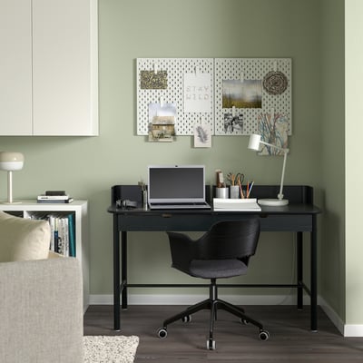 A cozy green home office with a black RIDSPÖ desk and chair, featuring integrated storage and a laptop. A pegboard with notes enhances the workspace.