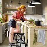 Woman photographs food in kitchen. Pink pants, red shirt, orange headband. Wooden counter, herbs, striped towels.