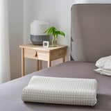 A modern bedroom a large bed with a ROSENSKÄRM pillow, next to a small wooden nightstand decorated with a unique lamp and a plant.