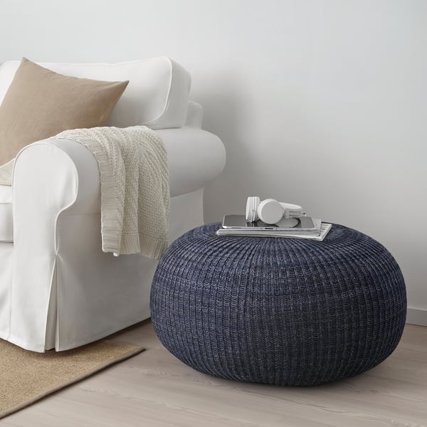 Dark blue knitted pouffe next to white chair. Round, soft, and textured. Small in size compared to chair.